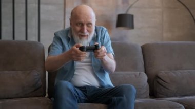 Zafer sevinci ile kablosuz kontrolörle video oyunu oynayan yaşlı bir adamın portresi. Kır saçlı emekli, koltukta oturan, duygusal olarak konsolda oynayan, oyunu kaybeden, mağlubiyet duygusu.