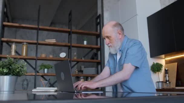 Зосереджений старий дідусь працює на ноутбуці зі свого домашнього офісу, друкуючи на клавіатурі ноутбука під час сидіння вдома на кухні. Свобода вдома, серфінг Інтернету, пошук інформації в Інтернеті. — стокове відео