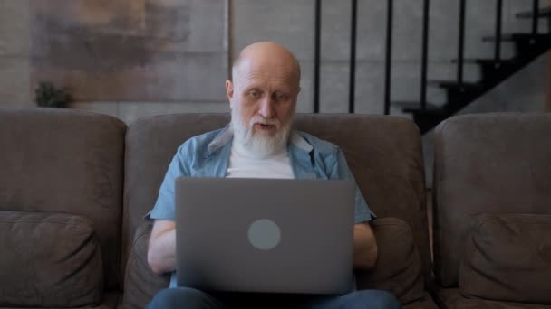高齢者の男性は、ビデオリンクを介して彼の家族に話す彼の手で笑顔とジェスチャーをズームソファの上に自宅に座ってラップトップ画面を見ています。老人はリモートで家に座って新しい職業を学ぶ. — ストック動画