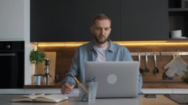 Tıraşsız erkek serbest çalışıyor. Evde dizüstü bilgisayarının başında oturuyor. Web kamerasıyla görüntülü sohbet yoluyla iletişim kuruyor. Öğrenci çevrimiçi bir öğretmenden öğrenir, konuşur ve not defterine metin yazar.