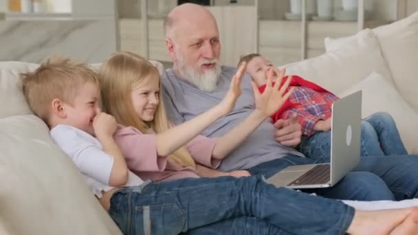 Gelukkige familie generaties, een oudere grijsharige grootvader met twee kleinkinderen en kleindochter, golf groet gebaar met hun handen naar webcam tijdens het communiceren via video chat zoom. — Stockvideo