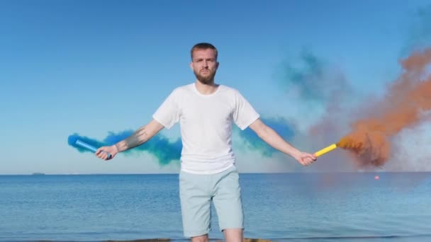 Молодий хлопець з бородою йде вздовж пляжу на тлі блакитного неба і океану, тримаючи феєрверк з кольоровим димом, кольоровими бомбами, димовими бомбами. Помаранчеві і сині димові бомби в руках людини . — стокове відео