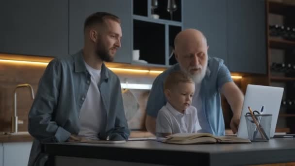 Happy family, father with a older grandfather and cute child using laptop, looking at webcam, waving his hands in greeting, communicating with relatives via zoom video chat while sitting at table. — Stock video