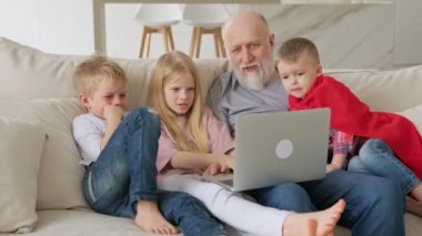 Mutlu aile nesilleri, iki torunu ve iki torunu olan yaşlı gri saçlı büyükbaba, video sohbet yoluyla akrabalarıyla iletişim kuruyor, evde otururken laptop ekranına bakan web kamerası.