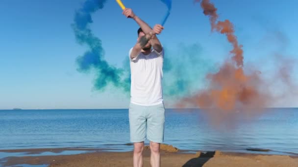 Jeune adolescent avec barbe se dresse sur fond de ciel bleu et océan tenant feu d'artifice fumée colorée, bombes colorées regarde la caméra. Bombes fumigènes orange et bleue dans les mains de l'homme, protestations de l'homme. — Video