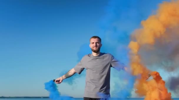 Młody facet z brodą spaceruje wzdłuż plaży na tle błękitnego nieba i oceanu, trzymając fajerwerki kolorowy dym, kolorowe bomby. Człowiek obraca się i bawi się niebiesko-pomarańczowym z bombą dymną na plaży. — Wideo stockowe