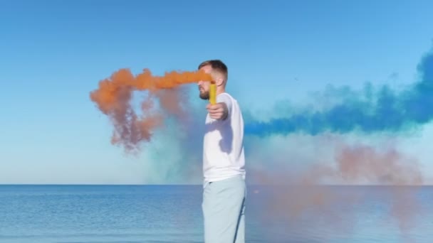 Junger Mann mit Bart läuft vor blauem Himmel und Meer am Strand entlang und hält Feuerwerkskörper, farbige Bomben. Mann dreht sich und spielt mit blauer und oranger Rauchbombe am Strand. — Stockvideo