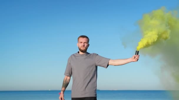 Молодий хлопець з бородою йде вздовж пляжу на тлі блакитного неба і океану, тримаючи жовтий феєрверк з кольоровим димом, кольоровими бомбами. Чоловік обертається жовтим димом сигнальна бомба з димовою бомбою на пляжі . — стокове відео