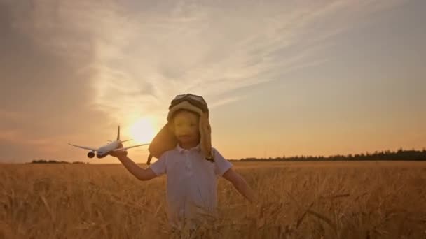 Au coucher du soleil, un garçon heureux au chapeau et aux lunettes de pilote d'aviation jouant avec un avion dans ses mains court sur un champ de blé de prairie. Rêves d'enfant devenir pilote d'aviation, joue avec l'avion jouet. — Video