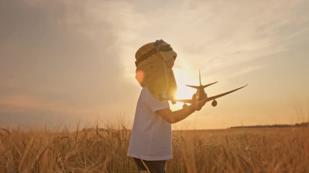 Slow-motion video, gelukkige jongen in hoed en luchtvaart piloot bril spelen met een vliegtuig in zijn handen bij zonsondergang op weide tarweveld. Kinderdromen die piloot worden, spelen met speelgoedvliegtuig. — Stockvideo
