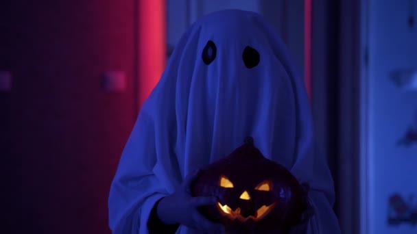 Enfant en costume de carnaval halloween fantôme en couverture blanche et avec citrouille rougeoyante dans ses mains se tient dans le couloir dans le clair de lune et orage chatoyant. Fantôme effrayant avec prise de gourde à la maison. — Video