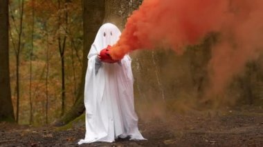 Cadılar Bayramı 'nda beyaz battaniyeyle hayalet kostümü giymiş ve ellerinde portakal dumanı tüttüren bir çocuk sonbahar ormanı içinde duruyor. Cadılar Bayramı 'nda balkabağı Jack' li ürkütücü hayalet..