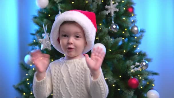 Mały chłopiec w białym swetrze i czerwony kapelusz w domu na tle Nowego Roku świecące fale choinki do kamery, rozmawia z rodziną w wideo czat i uśmiecha. — Wideo stockowe