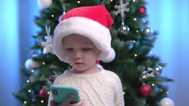 クリスマスツリーの波を背景に新年の赤い帽子で幸せな男の子と遠くの親戚とビデオチャットで話をします。男の子はクリスマスの挨拶を言います,オンライン携帯電話を介して通信. — ストック動画