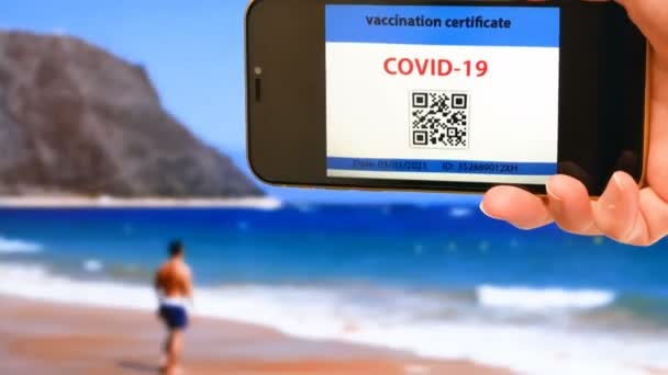 携帯電話での予防接種パスポートの映像です背景にある手と海のビーチでの旅行を可能にします コロナウイルスに対するワクチンCovid 免責事項又は健康パスポート — ストック動画