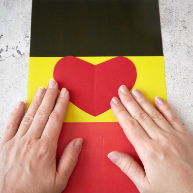 Almanya 'yı seviyorum. Eller, Almanya bayrağının arka planına kırmızı kağıt kalp koyuyor. Alman Birliği Günü - 3 Ekim, yakın plan