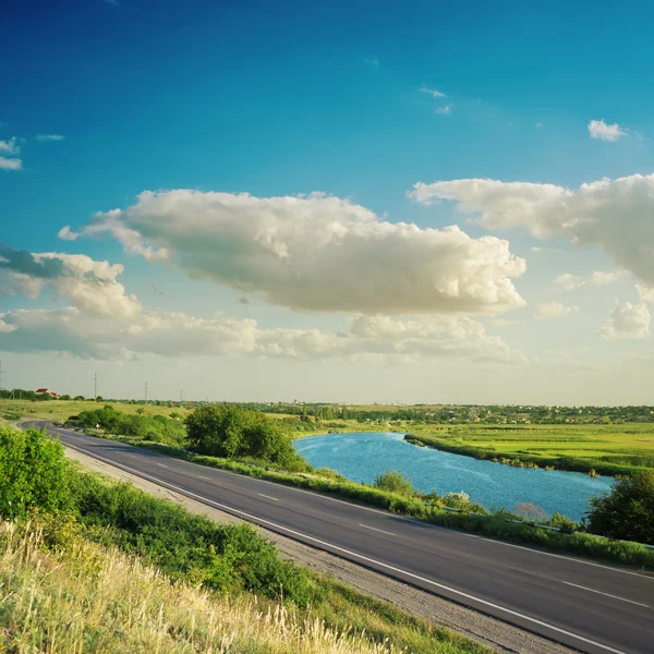 Road near river under clouds in blue sky on suset — Stock Photo, Image