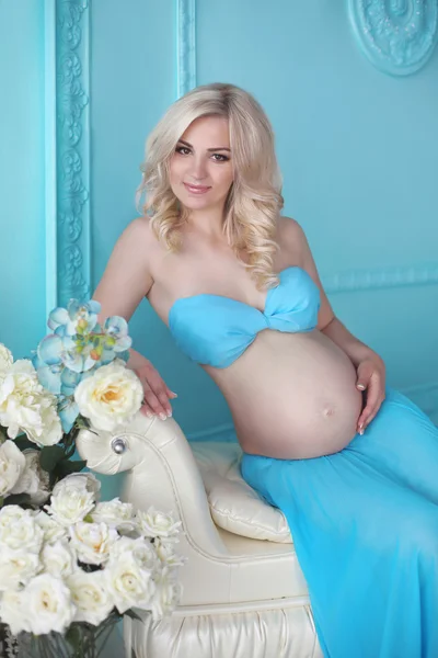 Atraktivní těhotná blondýnka dotýká její břicho rukama, pózuje v moderní modré interiérů domů. Nový život. Blondýnka čeká dítě. — Stock fotografie