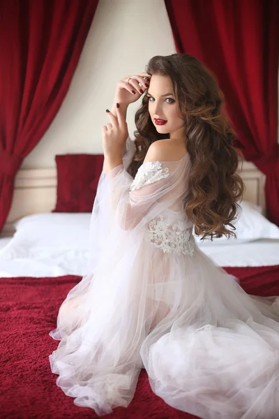 Güzel esmer gelin beyaz seksi boudoir sabahlık yatakta kırmızı ve beyaz yatak odasında poz giyiyor kıvırcık saç stili ile portre düğün. — Stok fotoğraf
