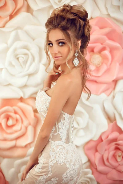 Attractive young bride with makeup and hairstyle, in wedding dress. Closeup portrait of young gorgeous woman over roses wall flowers. Studio shot.