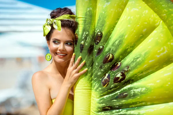 熱帯のビーチでインフレータブルキウイフルーツプールフロートマットレスで緑のイヤリングと美しいヒッピーの女の子の肖像画 夏休みの楽しみを持つ魅力的な幸せなブルネット — ストック写真