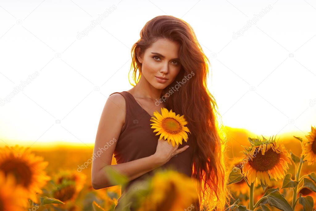 Beautiful young girl enjoying nature on the field of sunflowers at sunset. Summertime. Attractive brunette woman with long healthy hair.