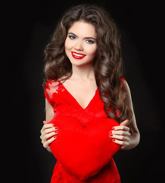 Belle fille souriante heureuse en robe rouge tenant le cœur de la Saint-Valentin. Brunette avec longue coiffure bouclée et lèvres rouges isolées sur fond noir . — Photo