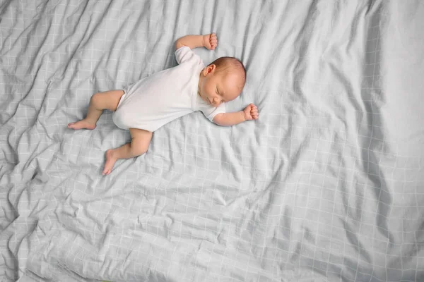 The tiny newborn is sleeping on the parent's bed. Place for text. Innocent Infant Boy in white bodysuit. Gray bedroom background. Childhood parenting. Sweet dream.Real life lifestyle small moments