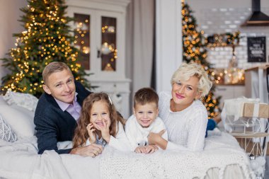 Klasik modern aile Noel 'i selamlıyor. Ebeveynler ve çocuklar yataktalar. Beyaz örülmüş kazaklar. Yeni yıl sabahı. İskandinav iç yatak odası bir çelenkle süslenmiş. Birlikte mutluluk