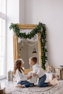 Kardeşler Noel Baba 'nın hediyelerini açar. Noel sabahı sıcak oturma odası. Hafif İskandinav iç mimarisi. Çocuklar beyaz kazaklar içinde neşeli mutludurlar. Çocukluk oyunu Neşeli kız çocuk