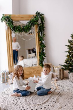 Kardeşler Noel Baba 'nın hediyelerini açar. Noel sabahı sıcak oturma odası. Hafif İskandinav iç mimarisi. Çocuklar beyaz kazaklar içinde neşeli mutludurlar. Çocukluk oyunu Neşeli kız çocuk
