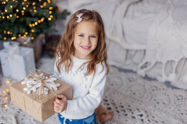  Çok güzel küçük bir kız elinde bir hediye tutuyor. Noel havası. Çocuk mutlu bir gülümseme ile hediye verir. Mesaj için yer. Mavi gözlü beyazlar gülümsüyor. Bokeh çelenkleri. Yeni Yıl