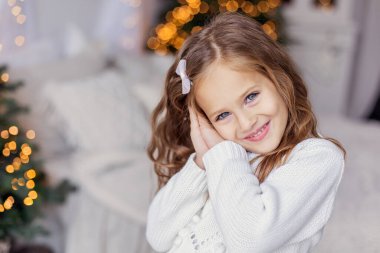 Güzel bir çocuk yeni yılda gülümser. Beyaz kazaklı, mavi gözlü, beyaz gözlü bir kız. Çok tatlı bebek gülüşü. Yakın plan portre. Yeni yıl ışıkları bokeh