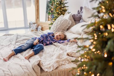 Yatakta yatan tatlı, güzel küçük kız. Çocuk Noel sabahını selamlıyor. Yatak odası yeni yıl için dekore edilmiştir. Eğlenceli çocukluk. Eve. Yaşam tarzının küçük anları. Samimi. Gülümseyen çocuk