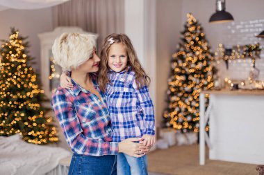 Anne ve kızı tişört giyerek sarılıyorlar. Günaydın Noel. Yeni yıl dekoru oturma odasında. Xmas havası. Hafta sonu tatili birlikteliği. Annelik çocukluğu. Beyaz kazak. Aile 