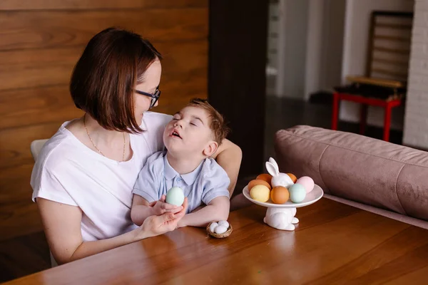 Mom and disabled son are painting Easter eggs in a lifestyle photo. child with cerebral palsy celebrates Easter at home. Happy family together. Inclusion. Kindness, mercy, care and happiness. autism