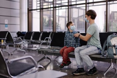 Havalimanında çocuğun koruyucu maskesini takan bir ebeveyn. Coronavirus salgınına gidin. Yeni normal yaşam tarzı. Baba ve oğul salonda kalkış için bekliyorlar. Güvenlik sosyal mesafesi