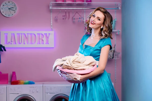 Beautiful woman in the laundry holds a basin with dirty clothes. The perfect housewife. Caricature exaggerated image of wife. Perfect makeup and hairstyle in 50s 60s pin-up style. Cleaning of clothes.