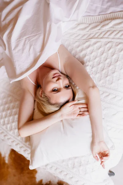 A young woman has slipped and lies in bed. White linens. Good morning, a great start to the day. Waking up in a good mood. Blonde with blue eyes. Healthy sleep comfortable bed
