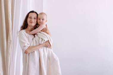 Anne beyaz arka planda bebeğe sarılıyor. Çevre ebeveynliği kavramı. Beyaz elbiseler ve bebek bezi doğal kumaşlar pamuklu kumaşlar. Anne sevgisi. Sarılma, gülümseme, mutluluk. Anne ve çocuk birlikteliği