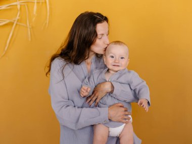 Gri elbiseli anne bir bebeği öpüyor. Anne ve çocuk. Anneliğin mutluluğu. Çevre vejetaryeni. Doğaya yakınlık. Mutlu çocukluk bakımı. Doğal kumaş. Sarı arkaplan kopyalama alanı