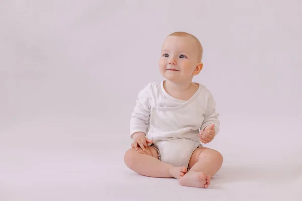 cute little kid in a white bodysuit sits on  background. 8 months old boy smiling.
