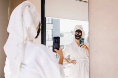 Kadının biri internette çamur maskesi takıyor. Güzellik salonunda selfie. Cep telefonu olan bir blogcu bir vlog kaydediyor. Neşeli ruh hali, kendini beğenmişlik. Siyah kille cilt bakımını rahatlatıyor. Eğlenceli. 