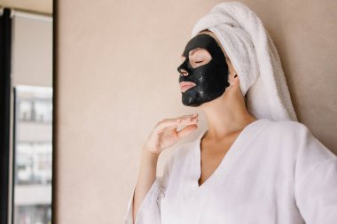 Killi anti-yaş maskeli kadın gülümsüyor. İyi ruh hali, pozitif kendini sevme. Kuaförde yüz siyaha boyanmış kaplıca tedavisi. Hafta sonu sabah rutini. Genç bir kızın canlı fotoğrafı. Cilt bakımı