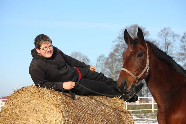 Ragazzo adolescente che riposa sulla balla di fieno e cavallo baia — Foto Stock