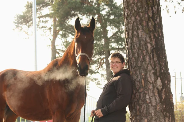 Мальчик-подросток и коричневая лошадь стоят у дерева — стоковое фото