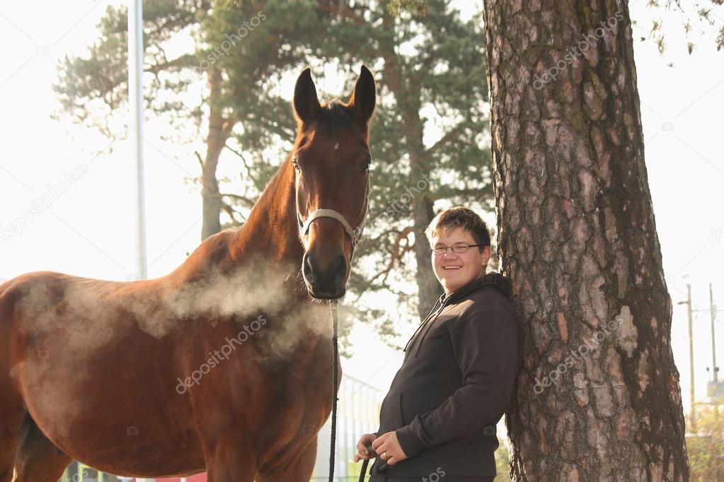 Teenager boy and brown horse standing near the tree