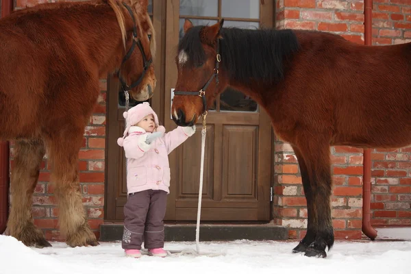 Little girl and two horses standing near the cottage door — Stock Photo, Image