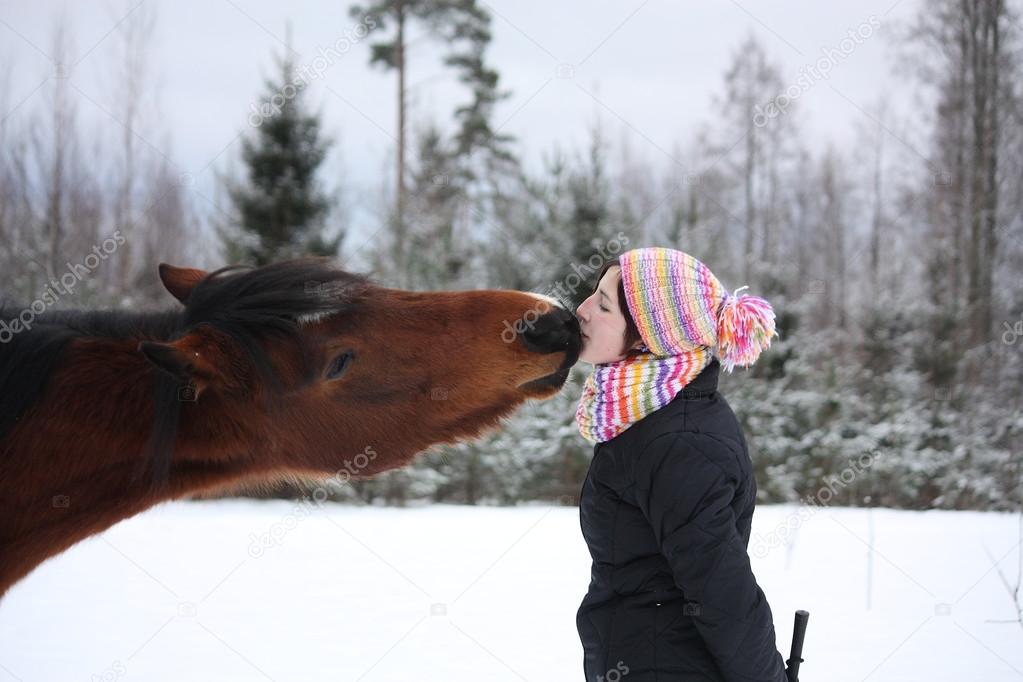 Beautiful teenager girl playfully kissing brown horse in winter