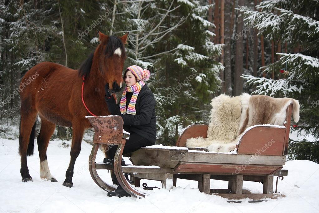 Teenager girl sitting in the sled with furs and brown horse 